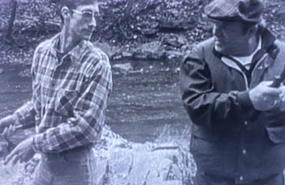 Two fishermen, one young, one old, stand in a creek comparing Blair Witch stories. 
