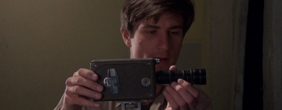 Robert De Niro plays Jon Rubin. Standing in a bare-walled room, Rubin, a light-skinned man with medium-long brown hair, is adjusting an analogue camera’s position on a tripod, eyes fixed on the lens.