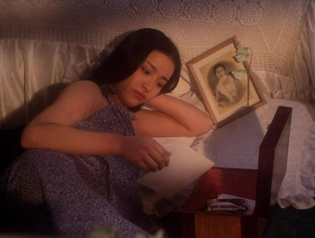 A young Asian woman laying in bed next to a framed photo, a flower, and a box of photos looking at a photo from the box.