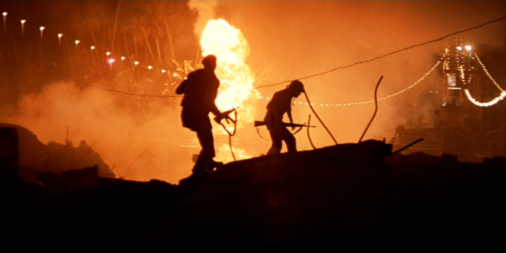 2 American soldiers holding machine gun rifles walk over a fiery hill by night