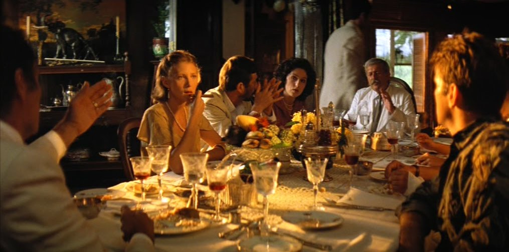 A group of French colonials sit in formal attire around a table with crystal goblets, white tablecloth, golden light.