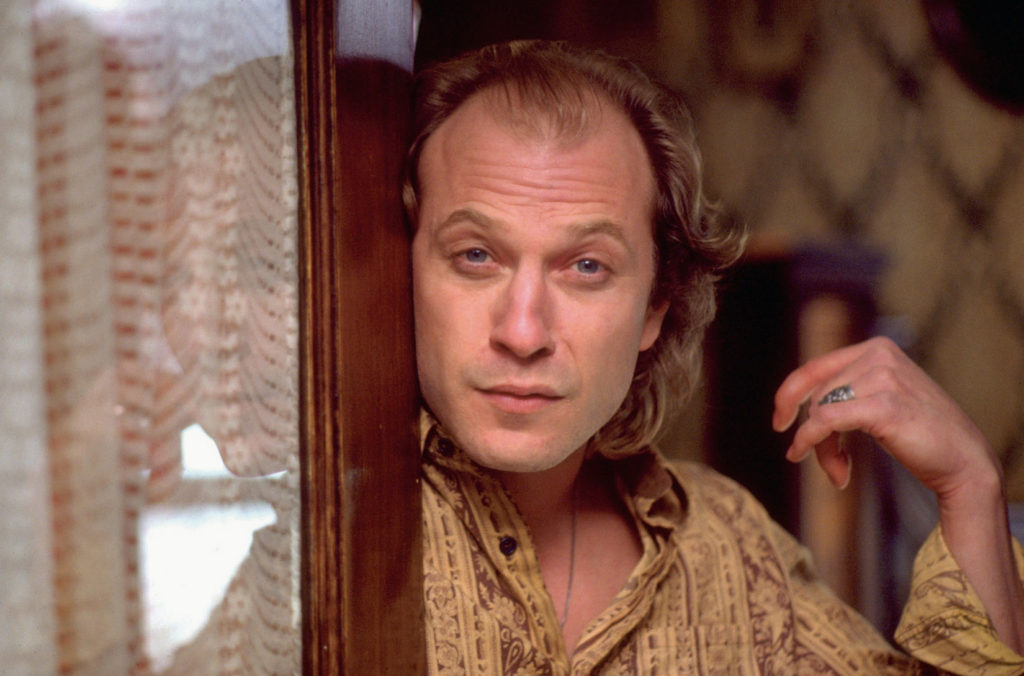 Buffalo Bill (played by Ted Levine) leaning against his front door, his face pressed against the door. He is wearing a yellow and brown shirt, and his left hand is curled inward toward his body.