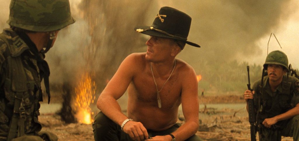 Robert Duvall as Colonel Kilgore kneels introspectively after napalm bombing on Vietnamese beach.