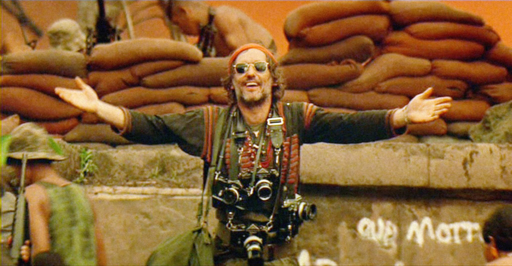 Dennis Hopper as Photojournalist with arms wide standing in front of ancient temple.
