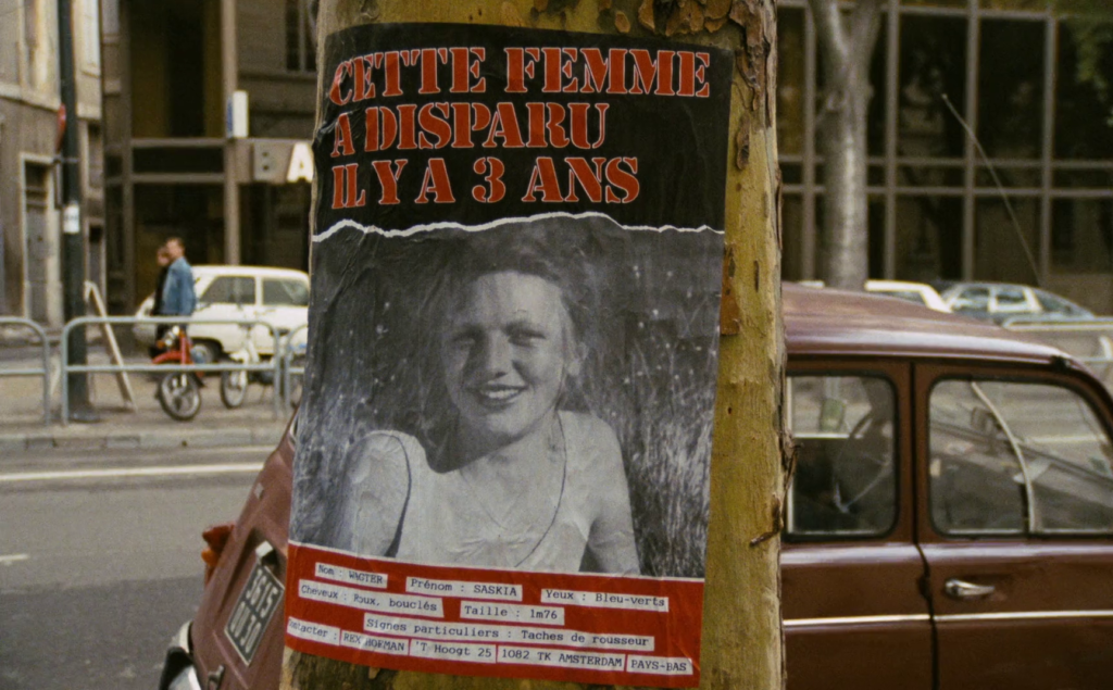 A close-up image of a missing person poster showing a black-and-white image of Saskia. The poster is glued to a tree on a city street