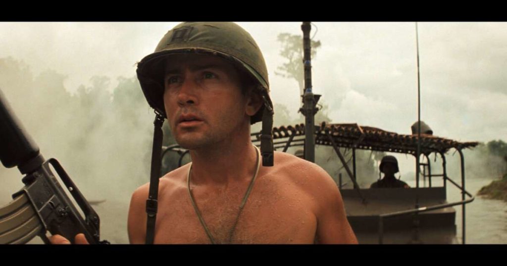 Martin Sheen as Captain Willard standing shirtless with M16 wearing helmet on bow of PBR.