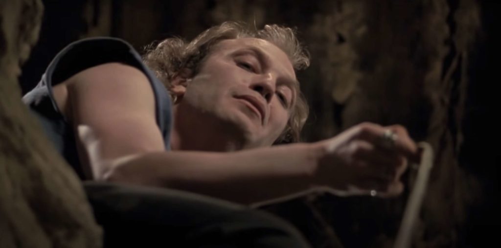 Buffalo Bill (played by Ted Levine), as seen from partway down the empty well in his basement. He is wearing a blue shirt. His hand is extended and holding a rope, as he makes an inscrutable expressio
