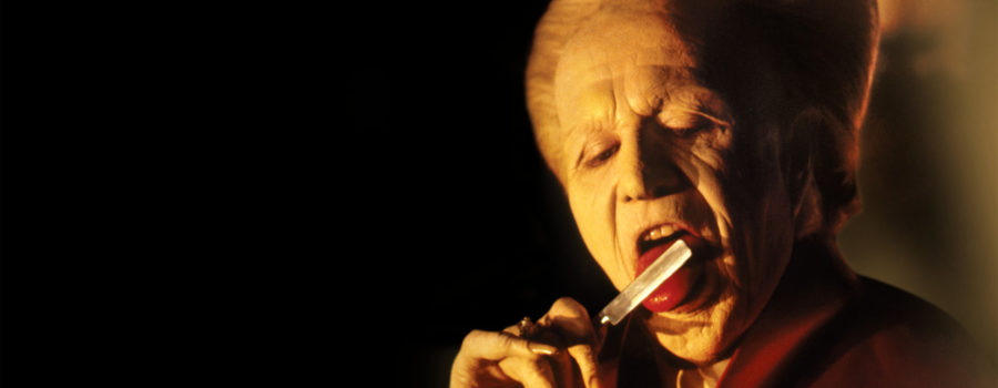 A menacing Gary Oldman as Count Dracula, wrinkled and pale, with his white and coiffed beehive hairdo, licks his razorblade to the right of the frame while shrouded in blackness.