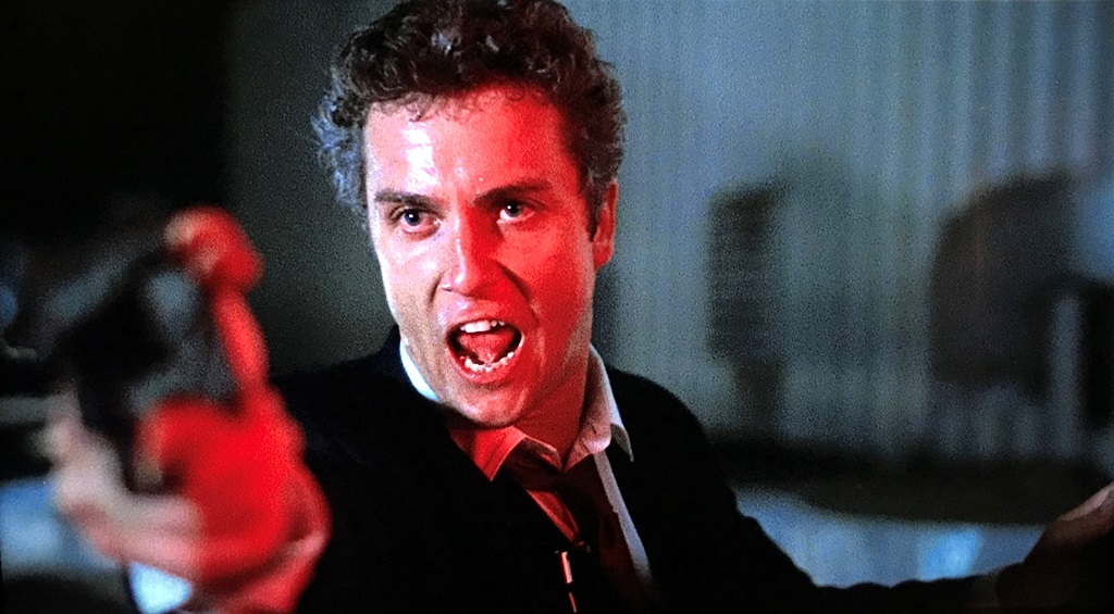 William L. Petersen as  Richard Chance in  To Live and Die in L.A. : Chance, a light-skinned adult man with dark, short wavy hair wearing a black suit is pointing a gun off camera. His facial expression is tense with his mouth wide open, and the right side of his face is illuminated by right red light.