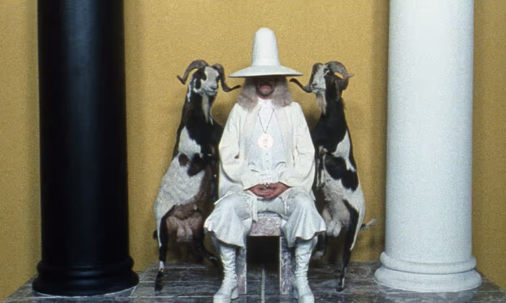 A man in a phallic white hat and white robes sits on a chair flanked by black and white goats. A black pillar is on his left, and a white pillar is on his right. The wall behind him is yellow.
