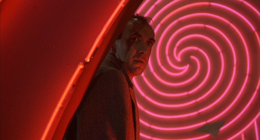 Sam Lowry (Jonathan Pryce) stands in front of a red neon sign, like the font that forms the title of the film.