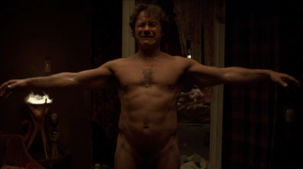 Harvey Keital as the Lieutenant, a light-skinned, adult man with short, curly dark hair in Bad Lieutenant. He is nude, wearing only a cross necklace, standing in the middle of a dimly-lit apartment room with his arms spread apart. He is crying.