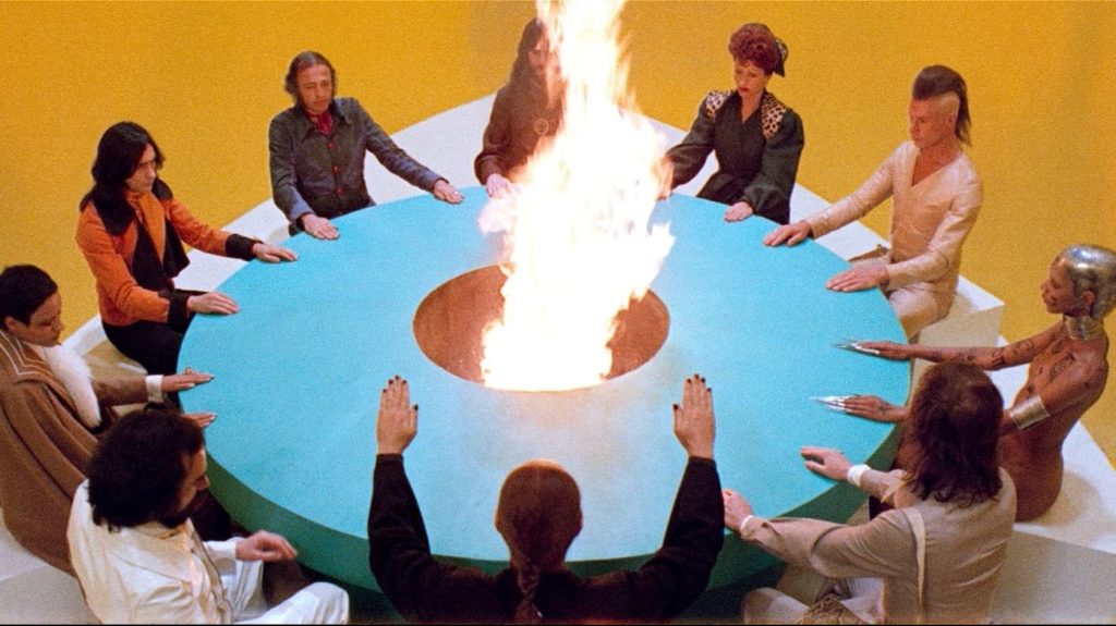 10 individuals dressed in bizarre clothing sit around a blue table with a hole spitting fire in the middle. They are sitting in a yellow room.