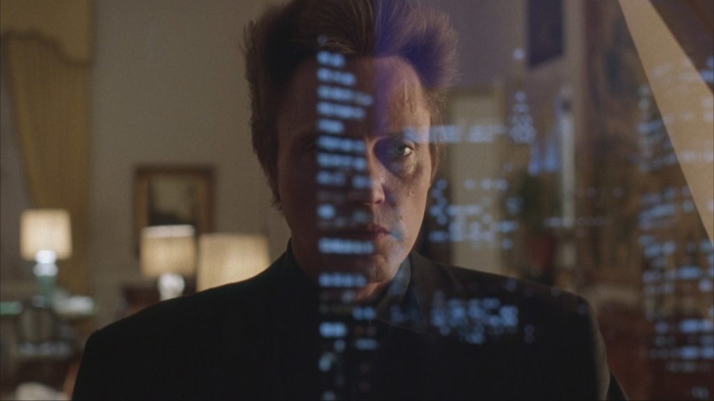 Christopher Walken as Frank White, an adult light-skinned man with blue eyes and spiked-up ash-blonde hair, is wearing an all-black suit. He is facing the camera standing in front of a window, and the skyline is reflected across his face on the window glass.