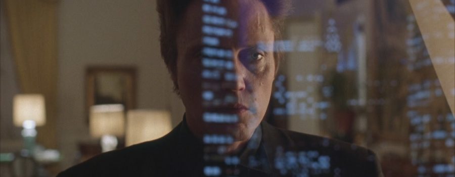Christopher Walken as Frank White, an adult light-skinned man with blue eyes and spiked-up ash-blonde hair, is wearing an all-black suit. He is facing the camera standing in front of a window, and the skyline is reflected across his face on the window glass.