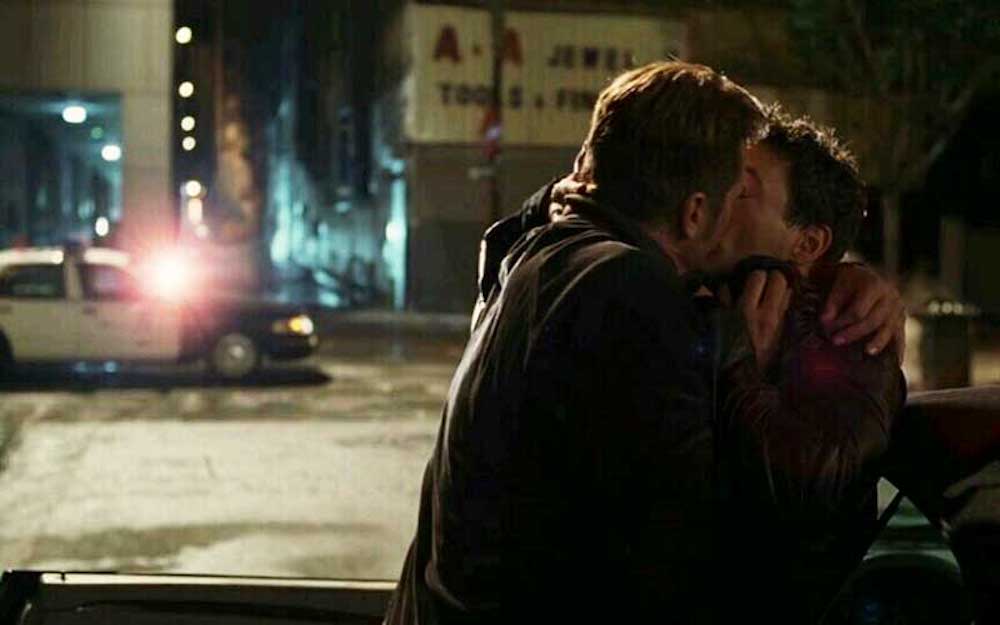 A light-brown-haired white man kissing a dark-brown-haired white man with a police car shining its light on them in the background.