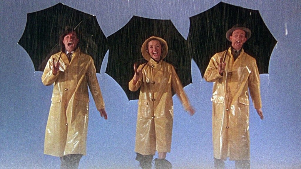A still from the opening scene of Singin’ in the Rain, with Gene Kelly, Debbie Reynolds, and Donald O’Connor in raincoats, holding umbrellas, walking through the rain
towards the camera