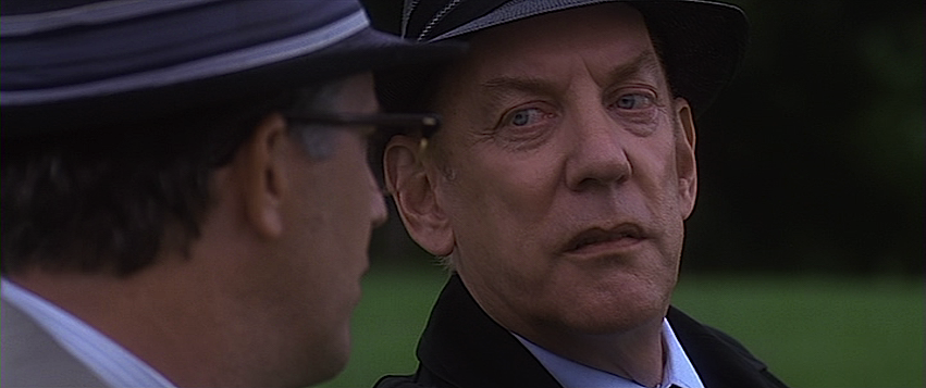 A shot of Donald Sutherland looking suspiciously towards Kevin Costner as they sit on a park bench, from the film JFK. 