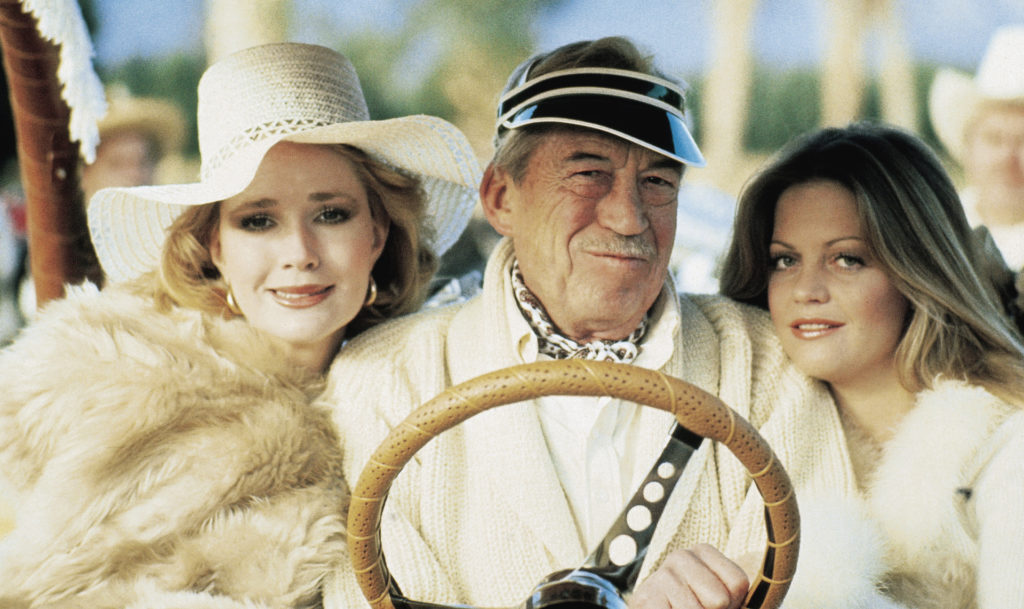 John Huston sitting in a golf cart between two much younger women, from the film Winter Kills. 