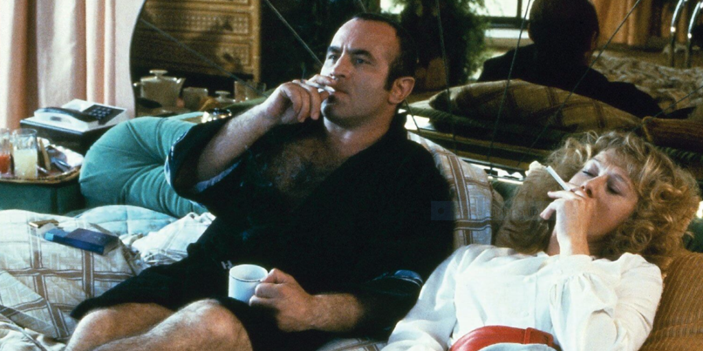 A man with an open black robe exposing his hairy chest is reclining on a bed, smoking a cigarette and holding a cup of coffee. There is a woman in a white dress lying to his right, also smoking. Their headboard is a reflective and gold, showing the back of the man's head.