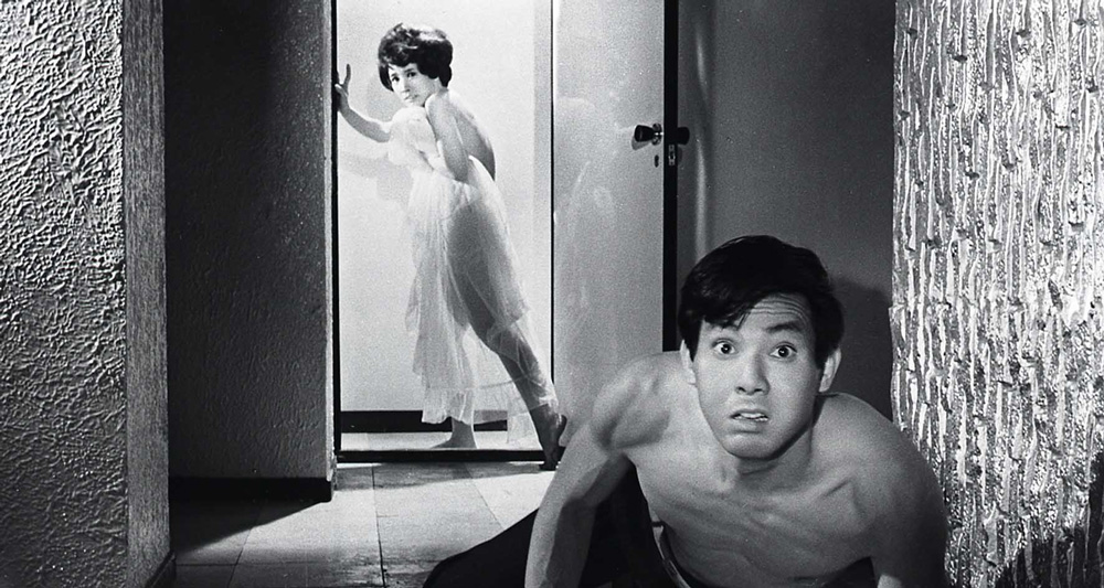 A naked woman draped in a thin robe is fleeing through a door in the background (while looking at the camera), while a topless man in the front stares intensely toward the camera.