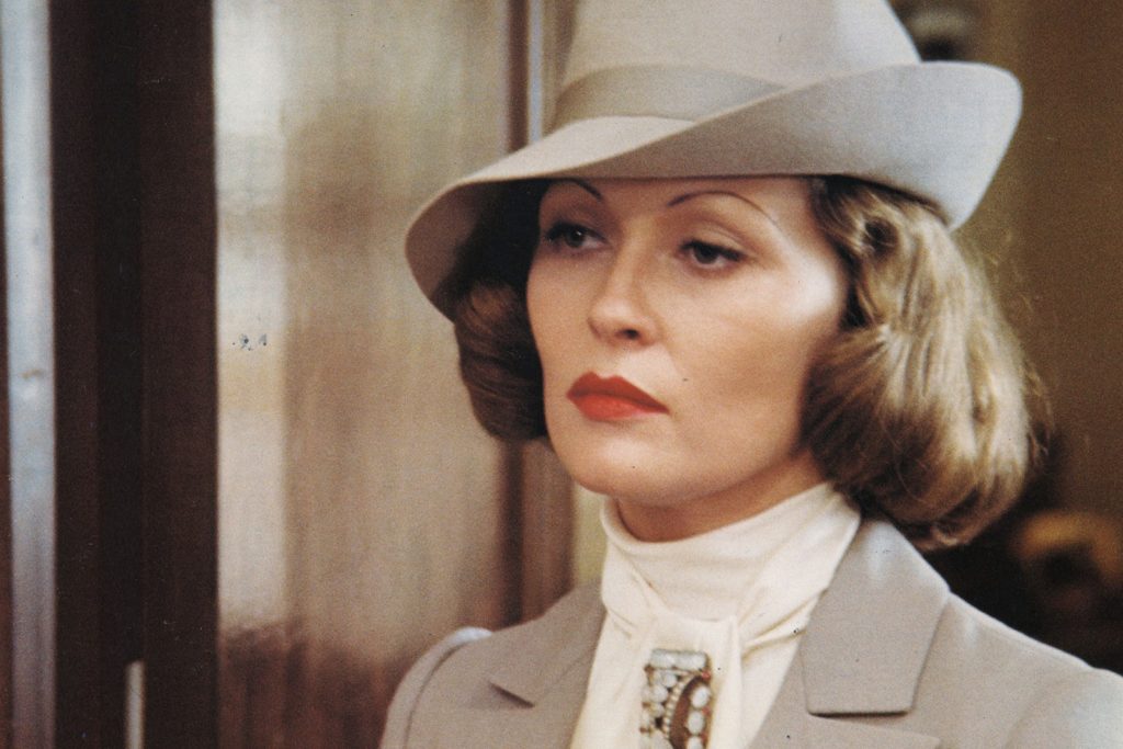 Faye Dunaway in character as Evelyn Mulwray in Chinatown. Evelyn is a light-skinned woman with curly chin-long hair and thin eyebrows. She looks into the distance unimpressed, wearing a grey dress suit and hat. 