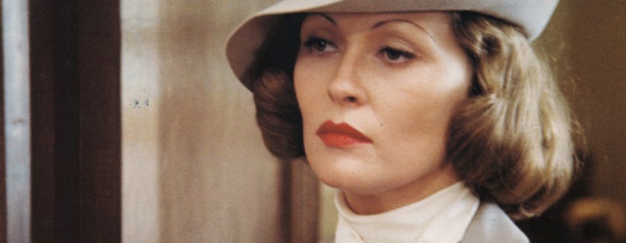 Faye Dunaway in character as Evelyn Mulwray in Chinatown. Evelyn is a light-skinned woman with curly chin-long hair and thin eyebrows. She looks into the distance unimpressed, wearing a grey dress suit and hat.