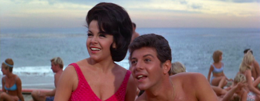 a white brunette woman sitting next to a white brunette man on a beach with many people in the background behind them