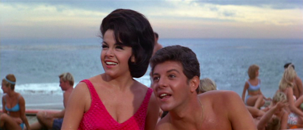 A white brunette woman sitting next to a white brunette man on a beach with many people in the background behind them