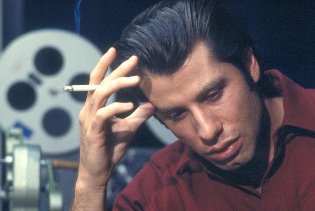 Medium close-up of a ponderous John Travolta (as soundman Jack Terry) in a maroon collared shirt leaning over with a cigarette in his right hand in the foreground, thumb pressed against the forehead. In the background are film reels and various editing equipment.