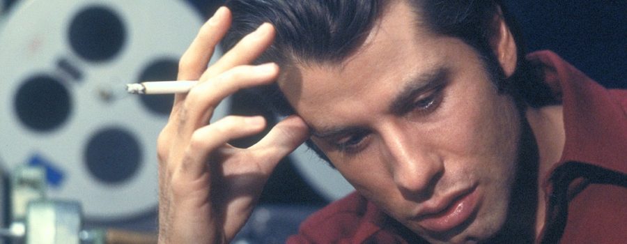 Medium close-up of a ponderous John Travolta (as soundman Jack Terry) in a maroon collared shirt leaning over with a cigarette in his right hand in the foreground, thumb pressed against the forehead. In the background are film reels and various editing equipment.