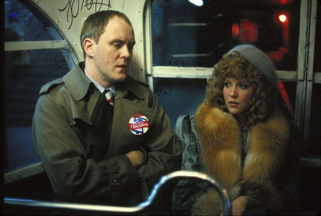 A medium two-shot featuring John Lithgow (as serial killer Burke) and Nancy Allen (as Sally) sitting close on a darkened, graffiti-speckled subway car. The balding Lithgow, in a dark green trenchcoat adorned with a 'Liberty Day' pin, has his arms are crossed with a menacing look on his face, aimed down slightly. Allen, in a silver and brown fur coat and with a knit cap atop her curly blond hair, looks into the distance, afraid but trying to remain calm.