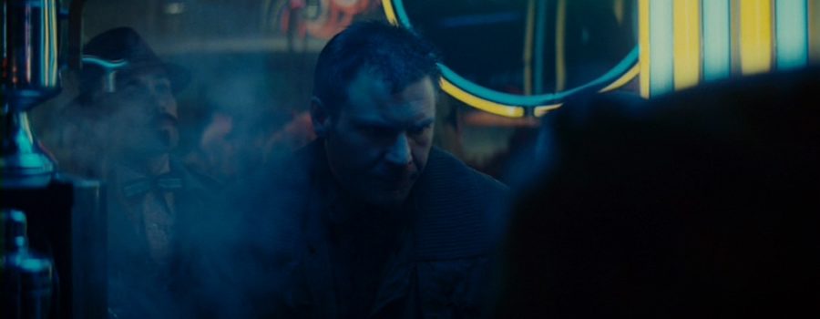 Deckard is sitting at a noodle restaurant, facing us, with his eyes turned downward. Behind him, Edward James Olmos’ Gaff stands menacingly.