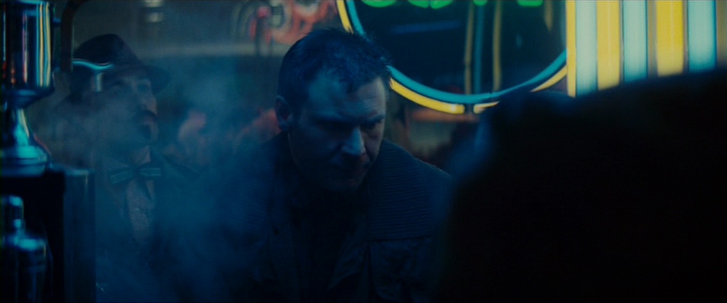 Deckard is sitting at a noodle restaurant, facing us, with his eyes turned downward. Behind him, Edward James Olmos’ Gaff stands menacingly.
