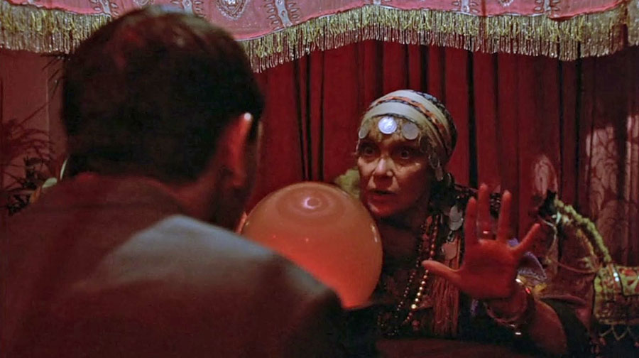 a fortune teller sits at a table with Pee Wee in a darkly lit room