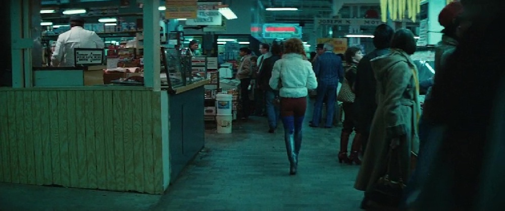 A back shot of Sally, wearing a puffy white jacket, high boots, and red-blue leggings, as she is walking through a crowded indoor marketplace.