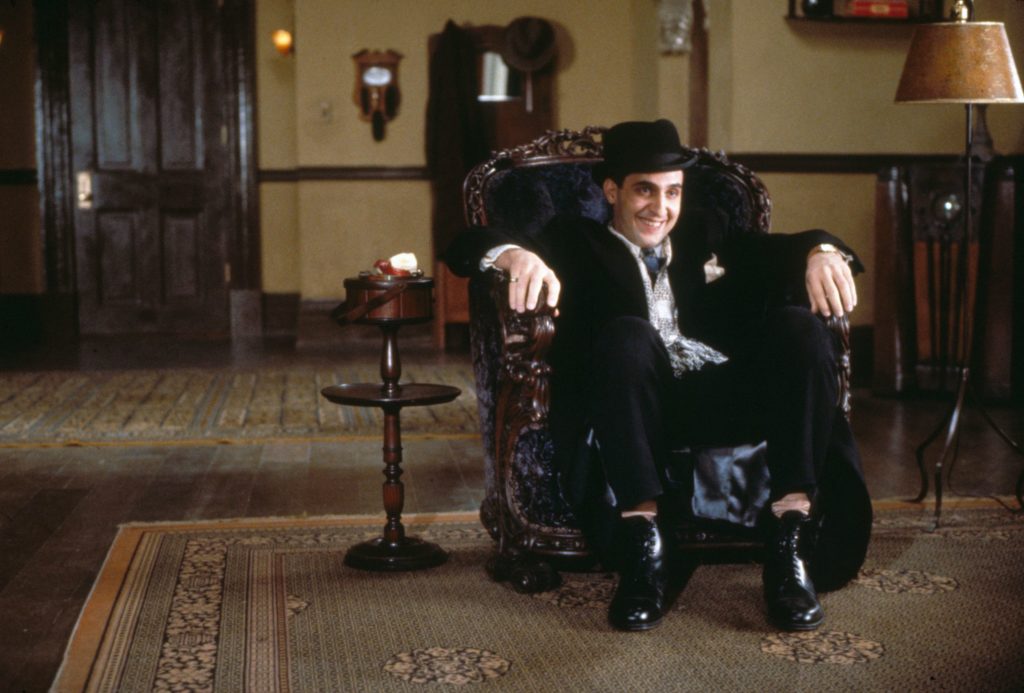 John Turturro as Bernie Bernbaum, a light-skinned, ark hair man in a dark suit and light shirt sitting in an ornamental armchair in a large room. Bernie is smiling at his conversation partner, who is not depicted in the shot