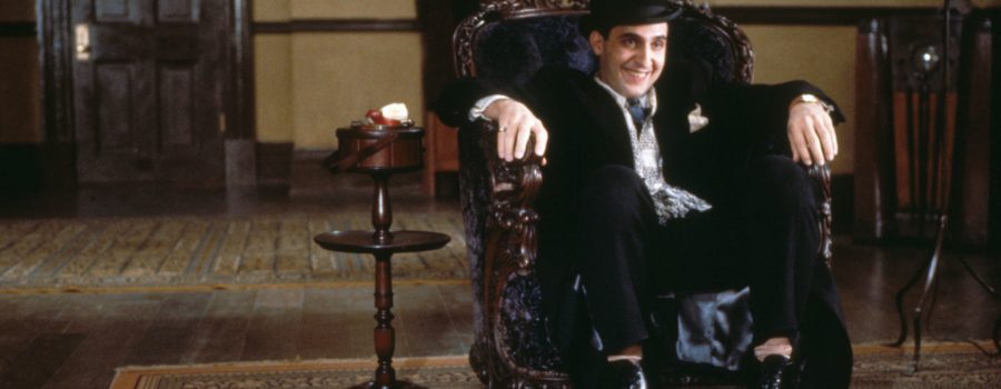 John Turturro as Bernie Bernbaum, a light-skinned, ark hair man in a dark suit and light shirt sitting in an ornamental armchair in a large room. Bernie is smiling at his conversation partner, who is not depicted in the shot