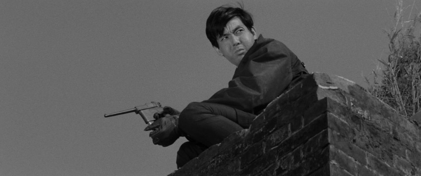 A man with dark hair dressed in dark clothing and leather gloves holds a gun while crouching on a rooftop, looking to the right side of the frame