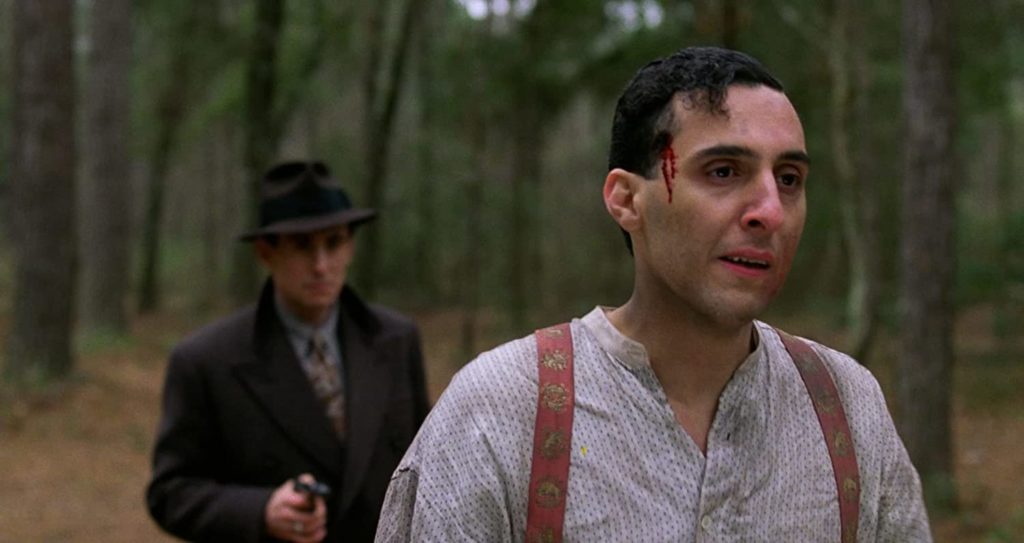 Gabriel Byrne as Tom points a gun to the back of John Turturro as Bernie Bernbaum in a forest setting.