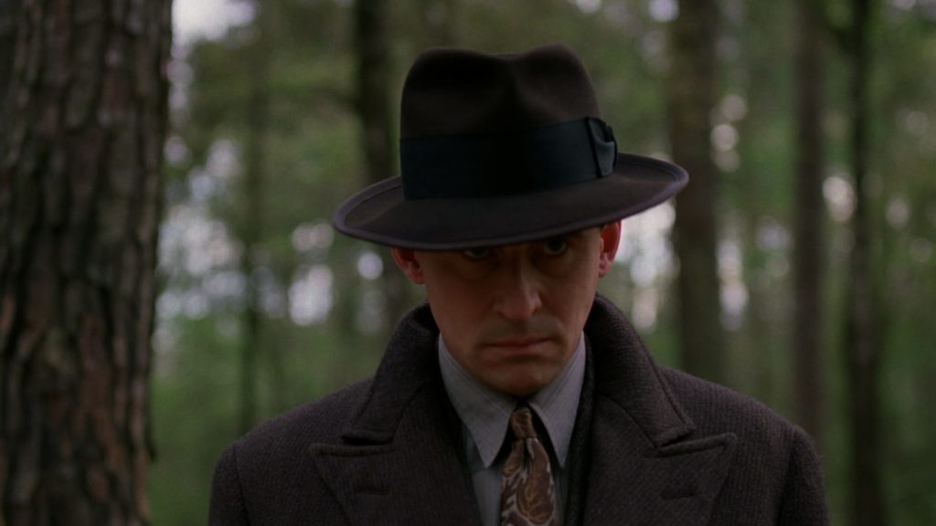 Gabriel Byrne as Tom with hat firmly on head standing in a forest.