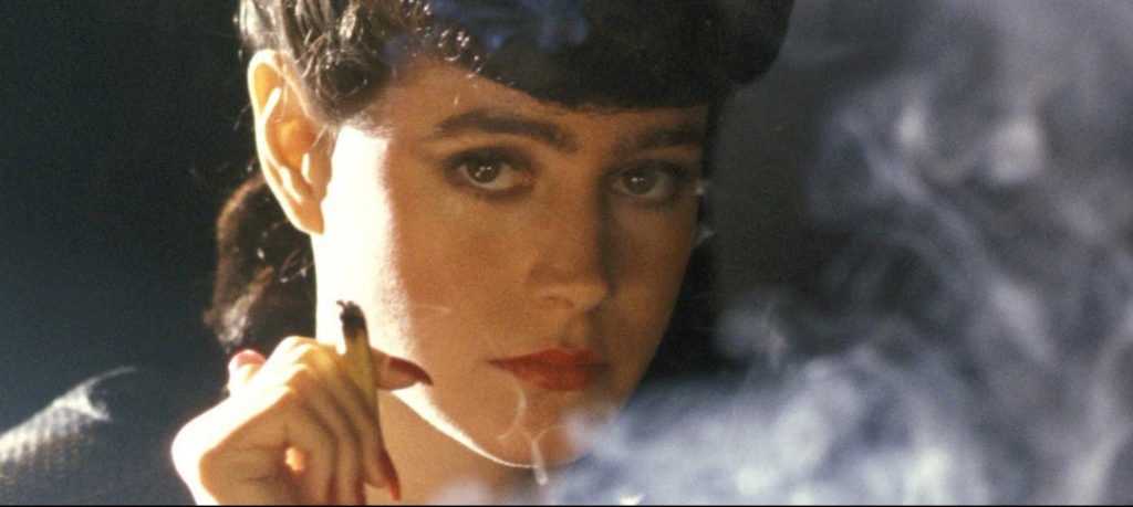 Sean Young as Rachael, a light-skinned woman with dark, made-up hair, wearing bright-red lipstick and nail polish, is encircled in a cloud of smoke from the cigarette she holds in her fingers while gazing into the camera.