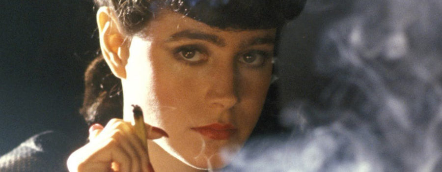 Sean Young as Rachael, a light-skinned woman with dark, made-up hair, wearing bright-red lipstick and nail polish, is encircled in a cloud of smoke from the cigarette she holds in her fingers while gazing into the camera.