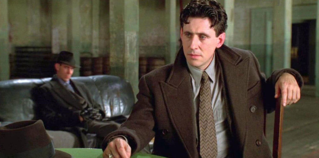 Gabriel Byrne as Tom Reagan,  light-skinned man with dark hair dressed in a brown coat, grey shirt, and brown zig-zag tie, is sitting on a chair in a warehouse. Blurry in the background is another man in a dark coat and hat, sitting on a black leather couch.