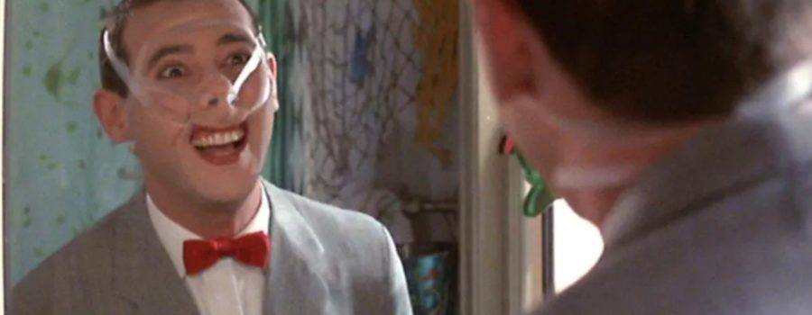 Paul Reubens as Pee Wee, a light-skinned man with short, light brown hair. He is shown from the back looking at himself in the mirror of a bathroom, wearing a face contraption the lifts his nose tip an upper lip.