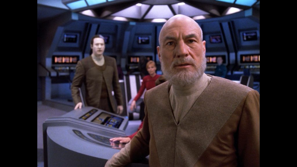 Patrick Stewart as Jean-Luc Picard stands on the bridge of the USS Pasteur looking surprised and confused. Gates McFadden as Captain Beverly Picard and Brent Spiner as Data watch in the background.