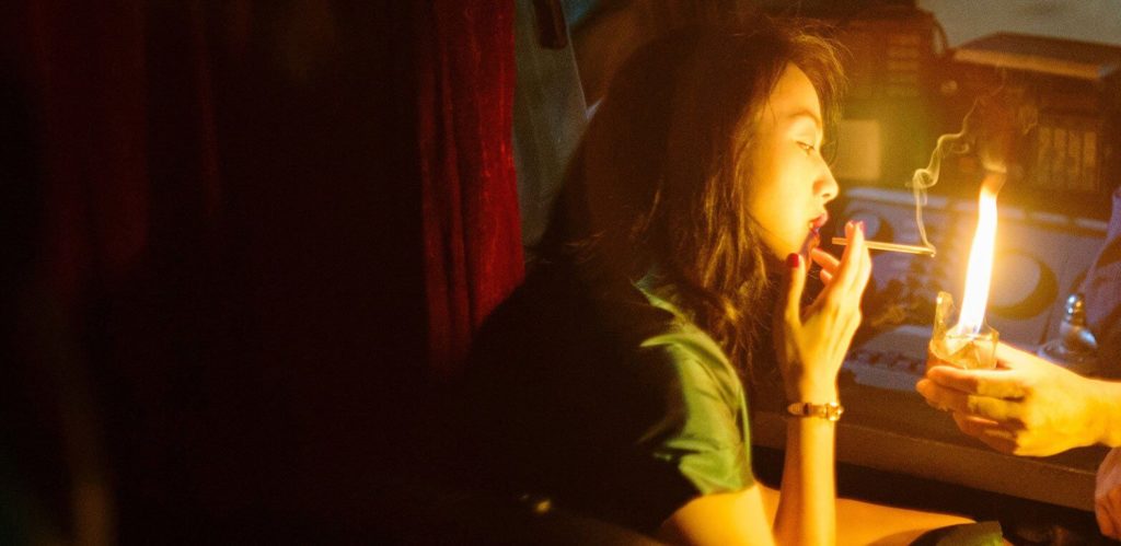 A woman in a green dress, played by actress Tang Wei, sits in a dark room and holds a lit cigarette, smoke billowing from its end, while a hand extends just out of frame with a lighter.