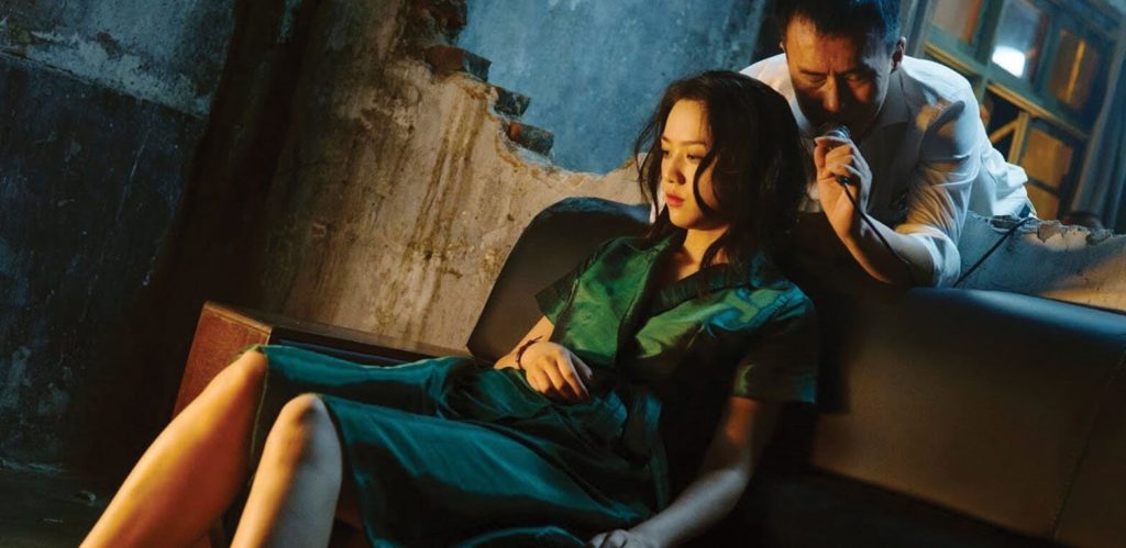 A woman in a green dress, played by actress Tang Wei, sits on a couch in a dilapidated house with half-demolished walls, vacantly looking downward at the ground. Through a half-demolished wall behind the couch, a man in a grey collared shirt, played by actor Huang Jue, leans over with closed eyes and reaches toward her, a hand close to her hair.