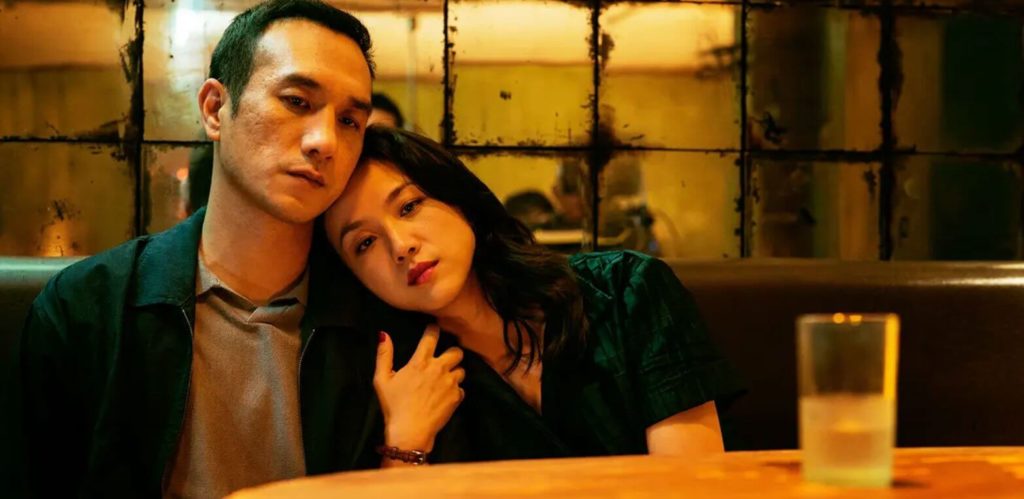 A woman in a green dress, played by actress Tang Wei, sits in a restaurant booth, her head leaned against the shoulder of a man in a green jacket and grey collared shirt, played by actor Huang Jue. The woman's right arm is wrapped around the man's left arm, and they are both staring vacantly off in the direction of the camera. On the table, a half-full glass of water sits.