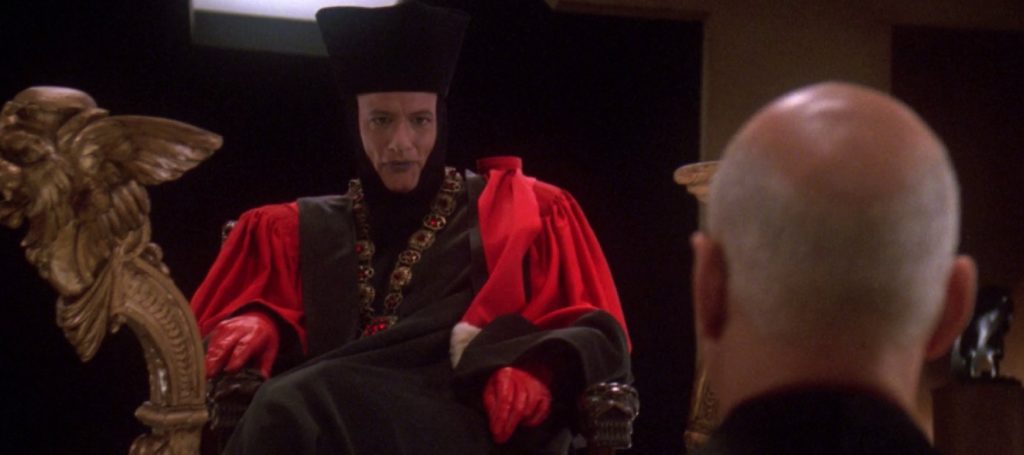 John de Lancie as Q dressed in gaudy red and black regal attire sits in golden throne. Patrick Stewart as  Captain Jean-Luc Picard stands facing Q with back to camera.
